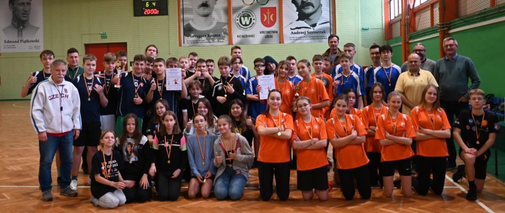 Duża grupa osób znajdująca się w sali gimnastycznej/sportowej pozują do wspólnego zdjęcia. Wszyscy mają na szyi medale oraz parę osób trzyma w ręce dyplomy wystąpienia w zawodach sportowych w piłkę ręczną.