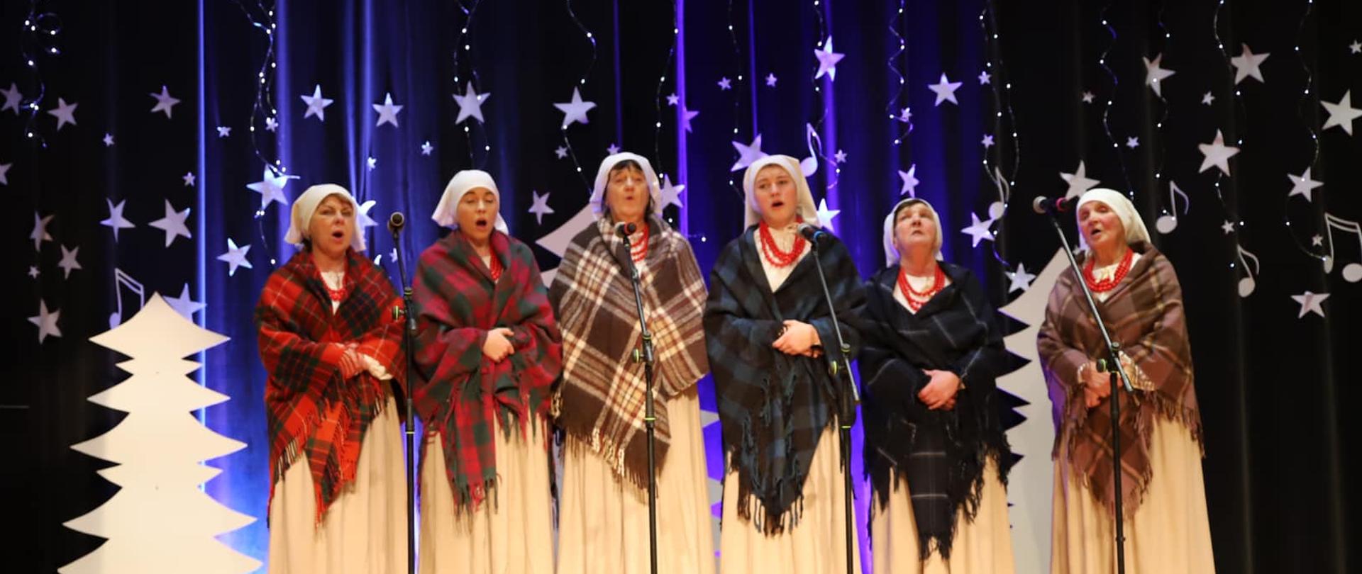 Zdjęcie przestawia stojącą na scenie grupę śpiewających kobiet, ubraną w wiejskie stroje z kocowymi chustami na ramionach i białymi chustkami na głowach. Nad śpiewającymi wisi baner z nazwą wydarzenia a z lewej strony stoi niebieski baner promocyjny Starostwa Powiatowego. Scena za występującym zespołem jest udekorowana w motywy świąteczne.