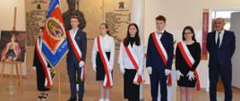 Poczet sztandarowy w budynku szkoły podstawowej przy tablicy pamiątkowej. Wszyscy uczniowie ubrani na galowo z biało czerwoną wstęgą.