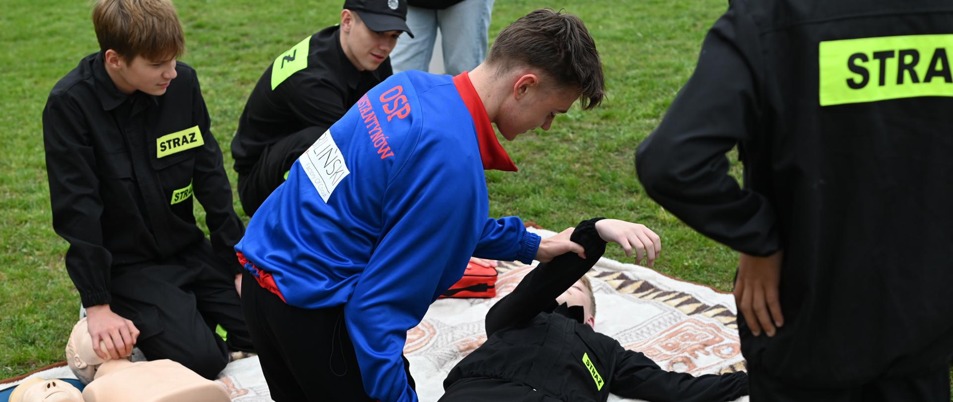 Młodzi strażacy podczas szkolenia z pierwszej pomocy na trawie. Jeden z uczestników ćwiczy ułożenie poszkodowanego w pozycji bezpiecznej, obok leżą fantomy do resuscytacji.