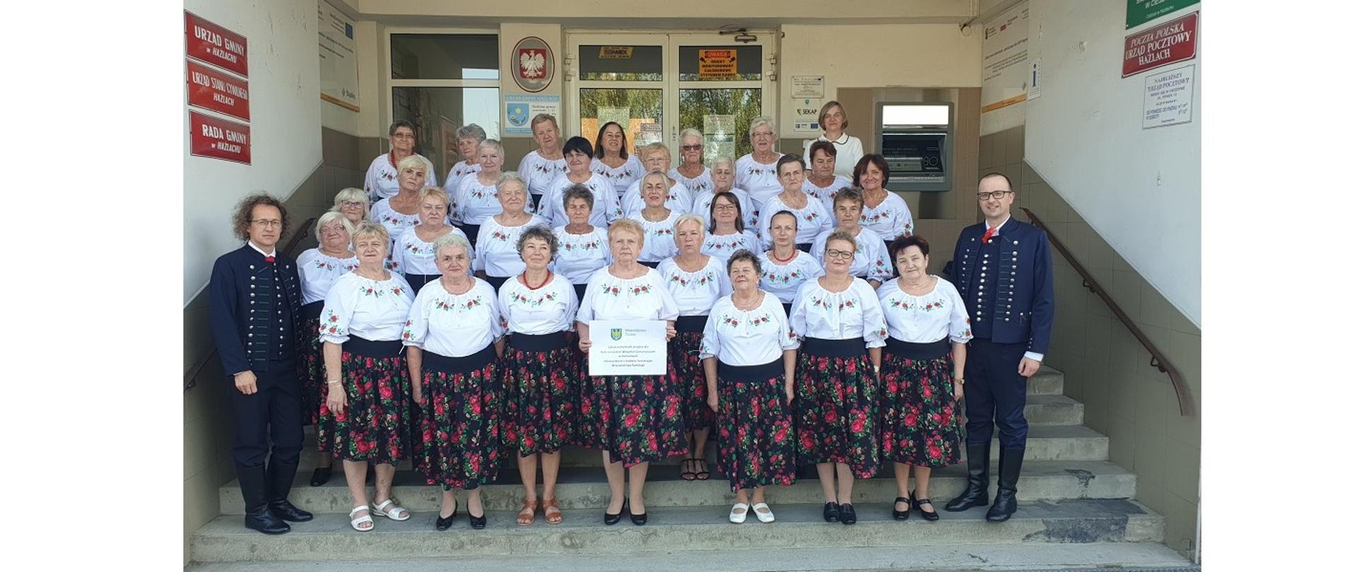 Członkinie Koła Gospodyń Wiejskich Zamarszczanka w nowych, jednolitych strojach przed wejściem do budynku Urzędu Gminy Hażlach. Towarzyszą im ubrani w męskie stroje cieszyńskie: Sołtys Zamarsk Klaudiusz Zawada i Wójt Gminy Hażlach Grzegorz Sikorski. Osoby pozują do fotografii stojąc amfiteatralnie w pięciu poziomych rzędach na stopniach schodów