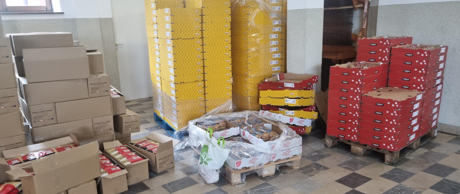 Cztery palety z kartonami oraz luzem ułożone pudełka na podłodze. Z paru kartonów wystają produkty żywnościowe takie jak pieczywo. 