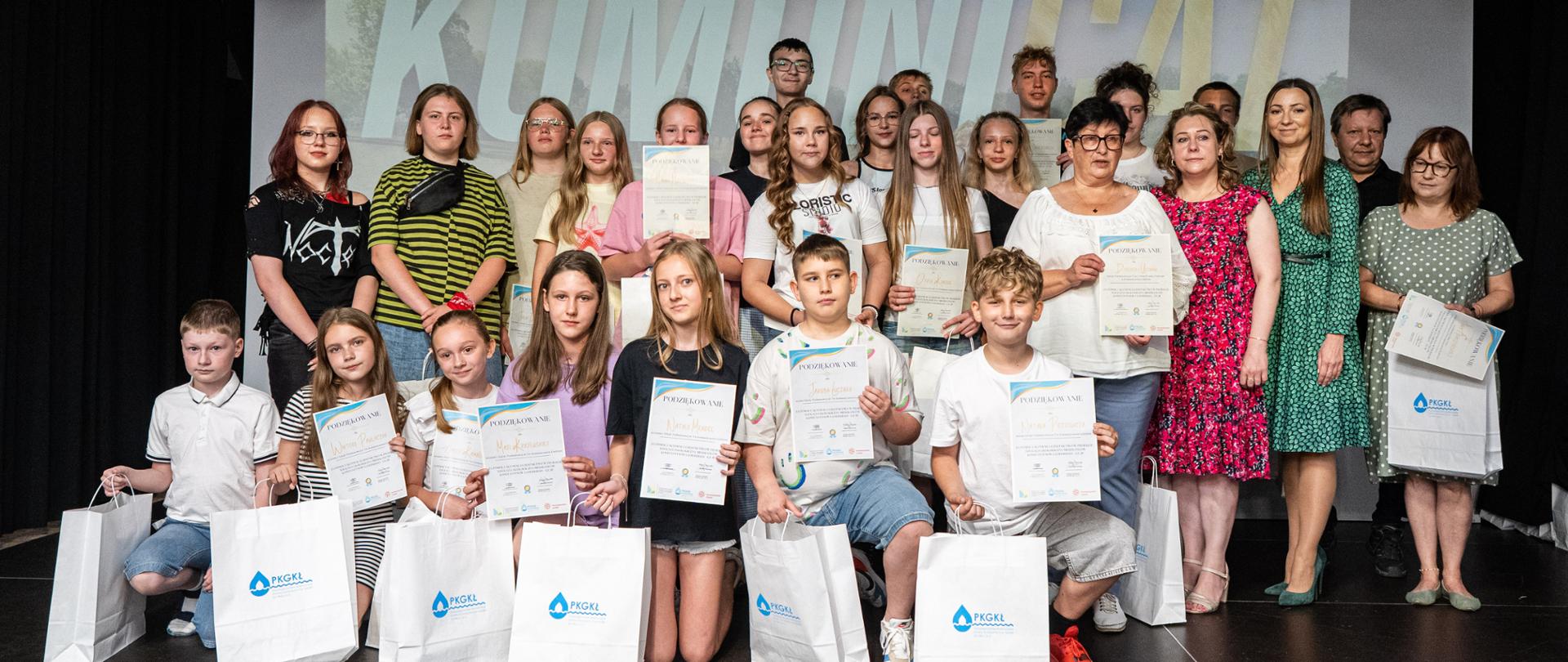 Grupa młodzieży oraz zaproszeni goście pozują na scenie z dyplomami i upominkami podczas spotkania podsumowującego projekt edukacyjny.