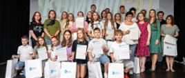 Grupa młodzieży oraz zaproszeni goście pozują na scenie z dyplomami i upominkami podczas spotkania podsumowującego projekt edukacyjny.