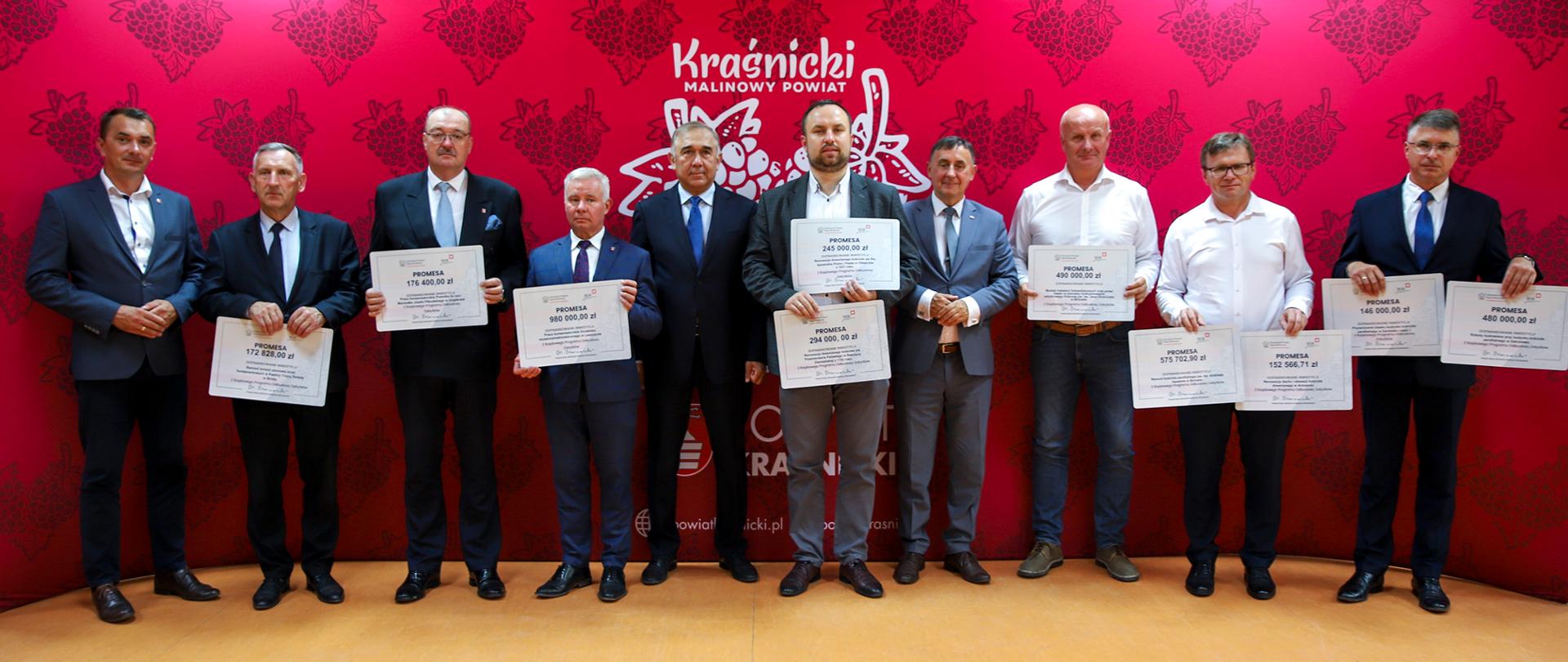 Zdjęcie przedstawia stojących w rzędzie ludzi, pozujących do pamiątkowego zdjęcia na tle czerwonej ścianki promocyjnej Powiatu Kraśnickiego. Uczestnicy trzymają w rękach promesy z przyznanymi kwotami.