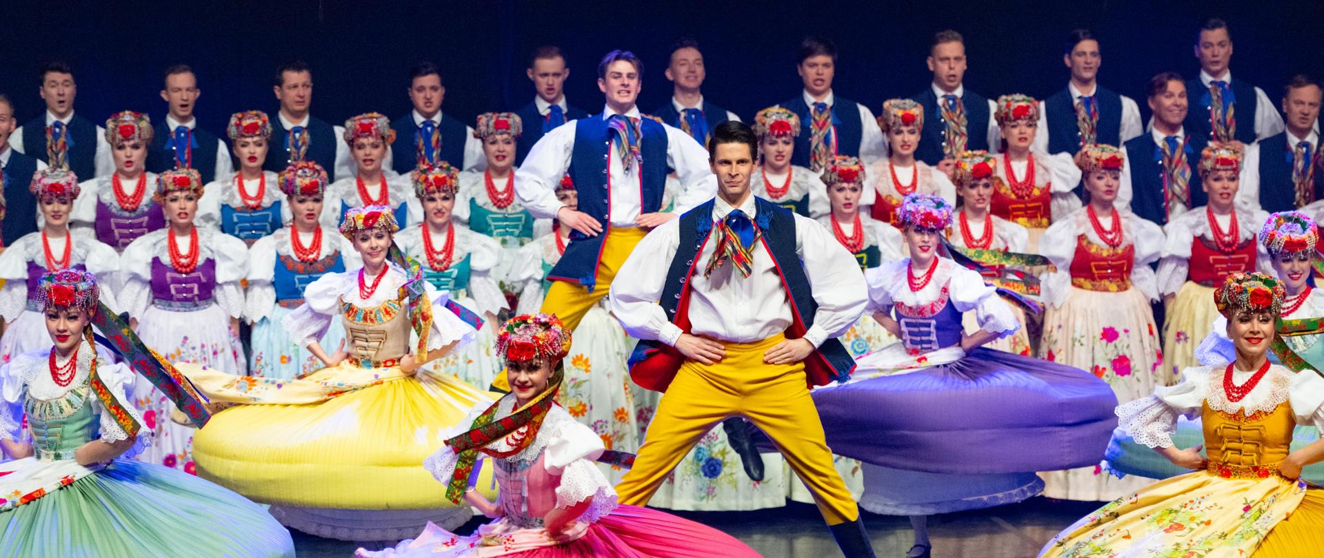 Tancerze w tradycyjnych strojach wykonują energiczny układ taneczny. Kobiety wirują w kolorowych, szerokich spódnicach, a mężczyzna w centrum sceny wykonuje skok.