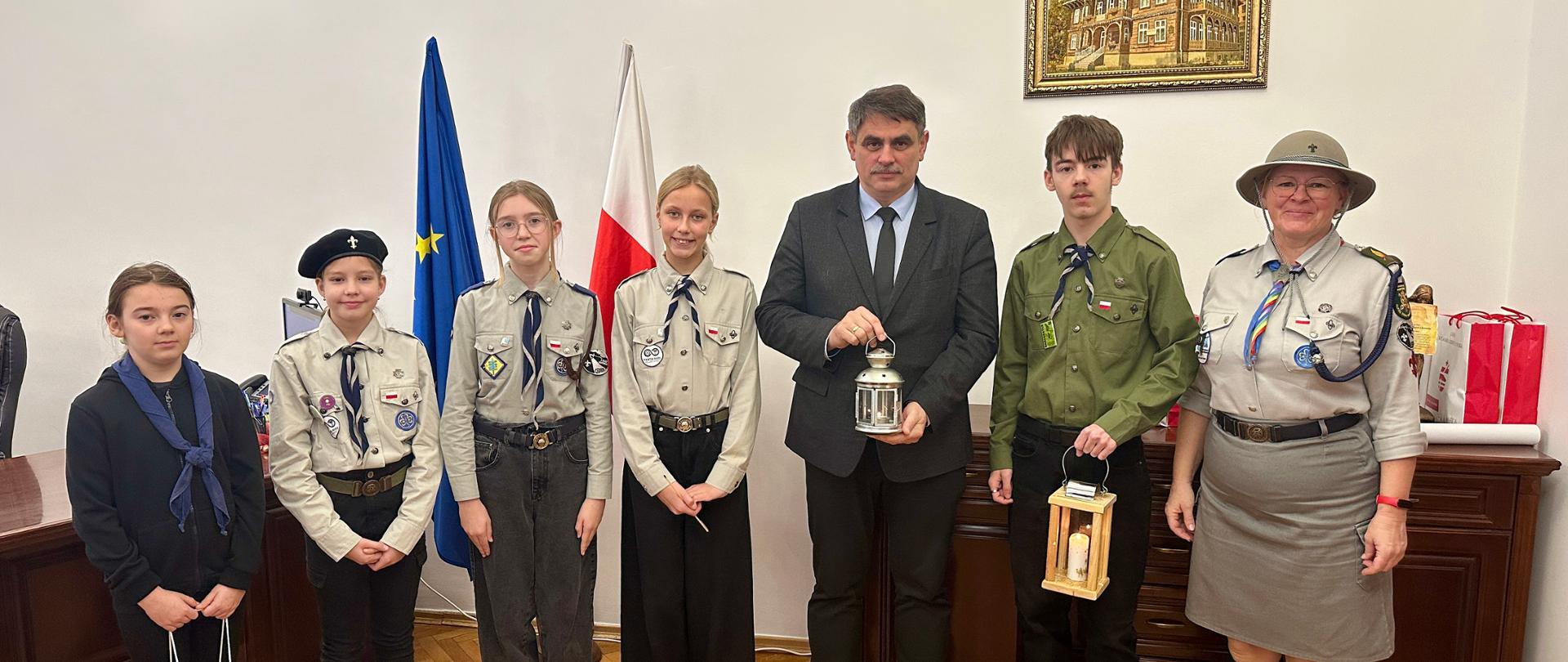 Grupa harcerzy i instruktorów w mundurach harcerskich stoi w gabinecie Urzędu Miasta. W centrum Burmistrz trzyma lampion z Betlejemskim Światłem Pokoju. W tle widoczne są flaga Polski, flaga Unii Europejskiej oraz godło państwowe na ścianie.