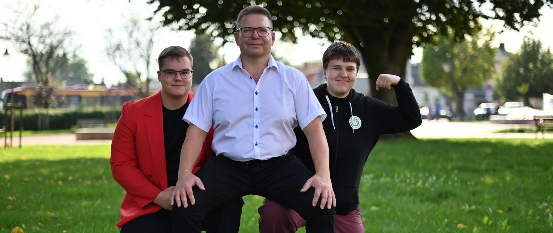 Trzy osoby klęczące w parku, w tle drzewo.