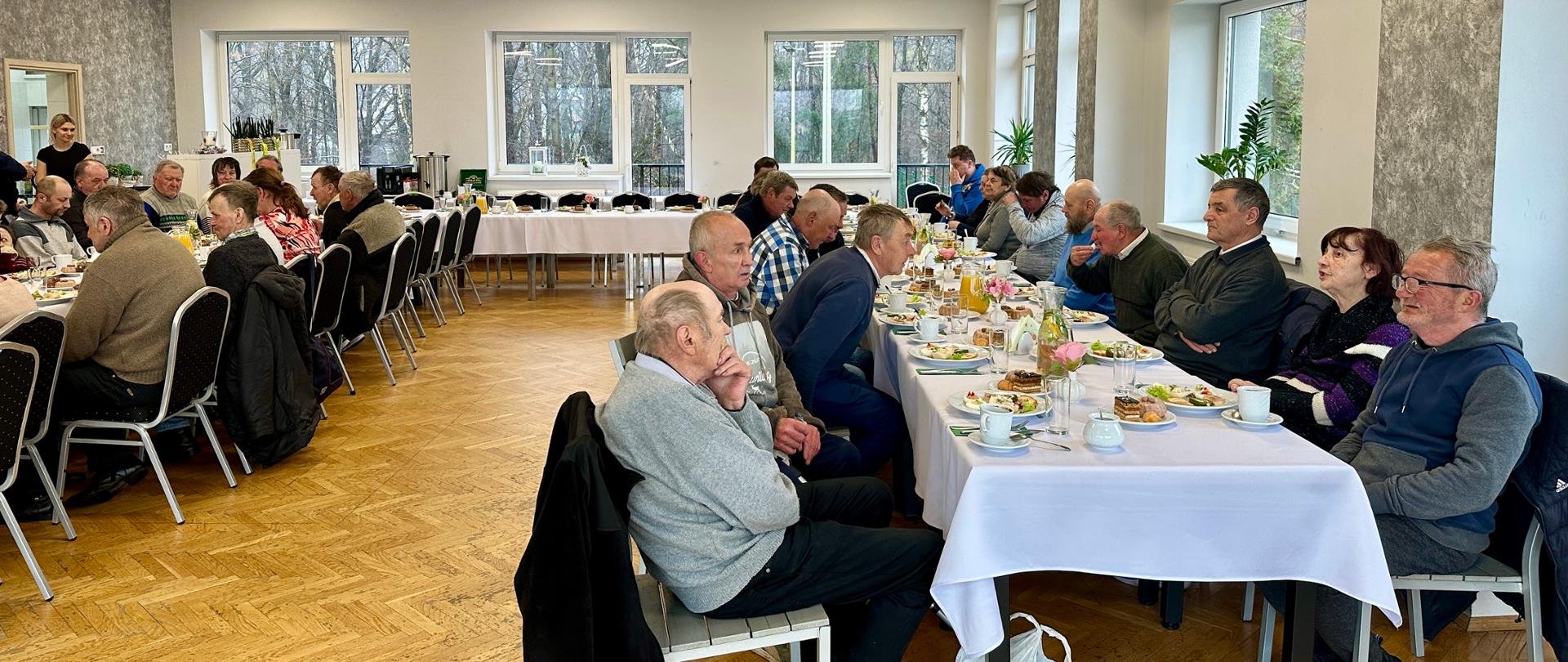 Jasna, przestronna sala, w której przy długich stołach nakrytych białymi obrusami siedzą uczestnicy wspólnego spotkania. Na stołach znajdują się posiłki, napoje i zastawa stołowa. W tle widać osoby rozmawiające oraz przygotowujące poczęstunek. Do wnętrza wpada naturalne światło z dużych okien, za którymi widoczne są drzewa. Atmosfera spokojna i integracyjna.