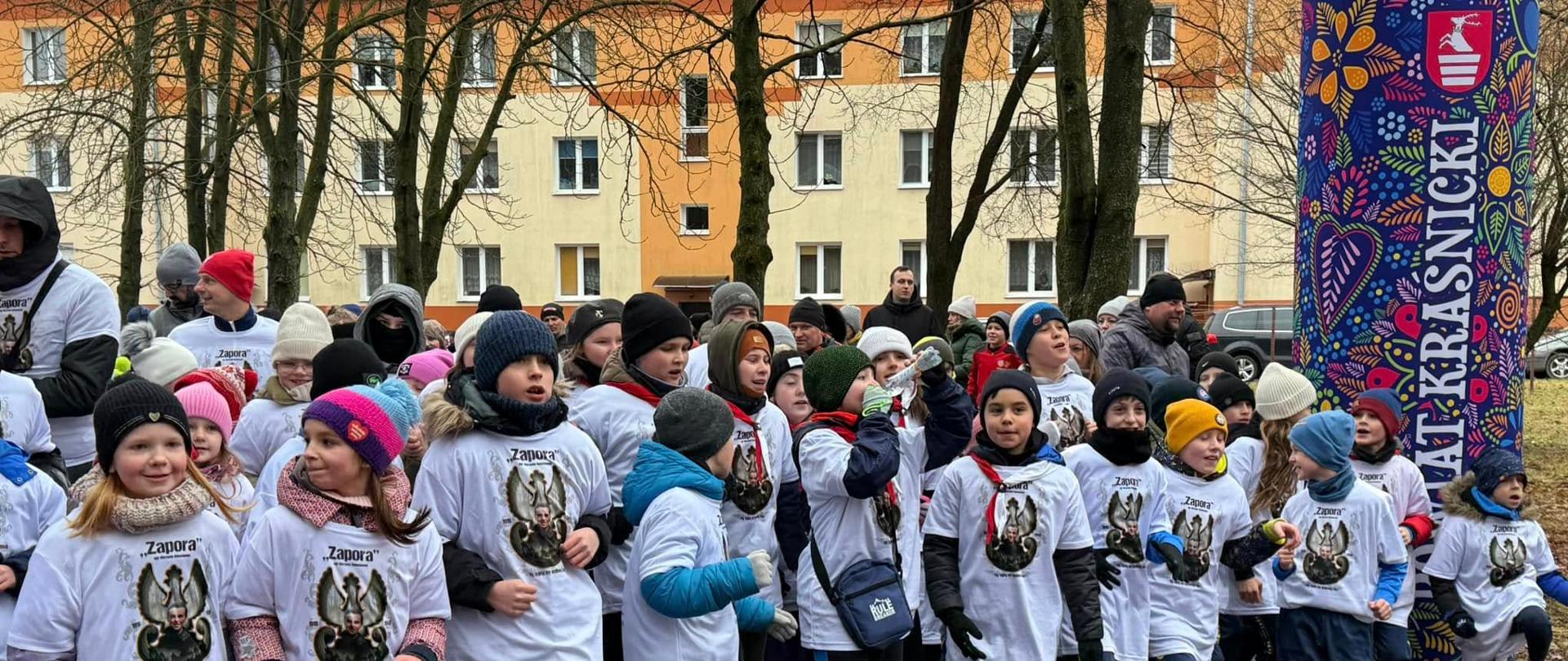 Na zdjęciu znajdują się uczestnicy biegu przygotowujący się do startu biegu. W większości są to dzieci, wszyscy są ubrani w białe koszulki z logotypem biegu. 