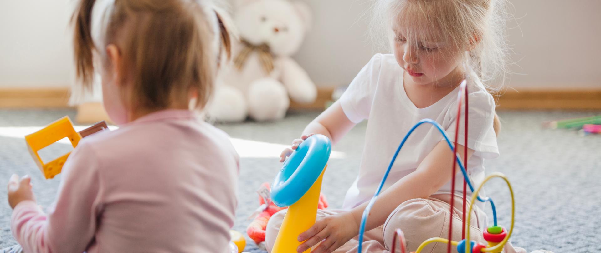 Dwie około trzyletnie dziewczynki siedzą na dywanie i bawią się zabawkami edukacyjnymi.
