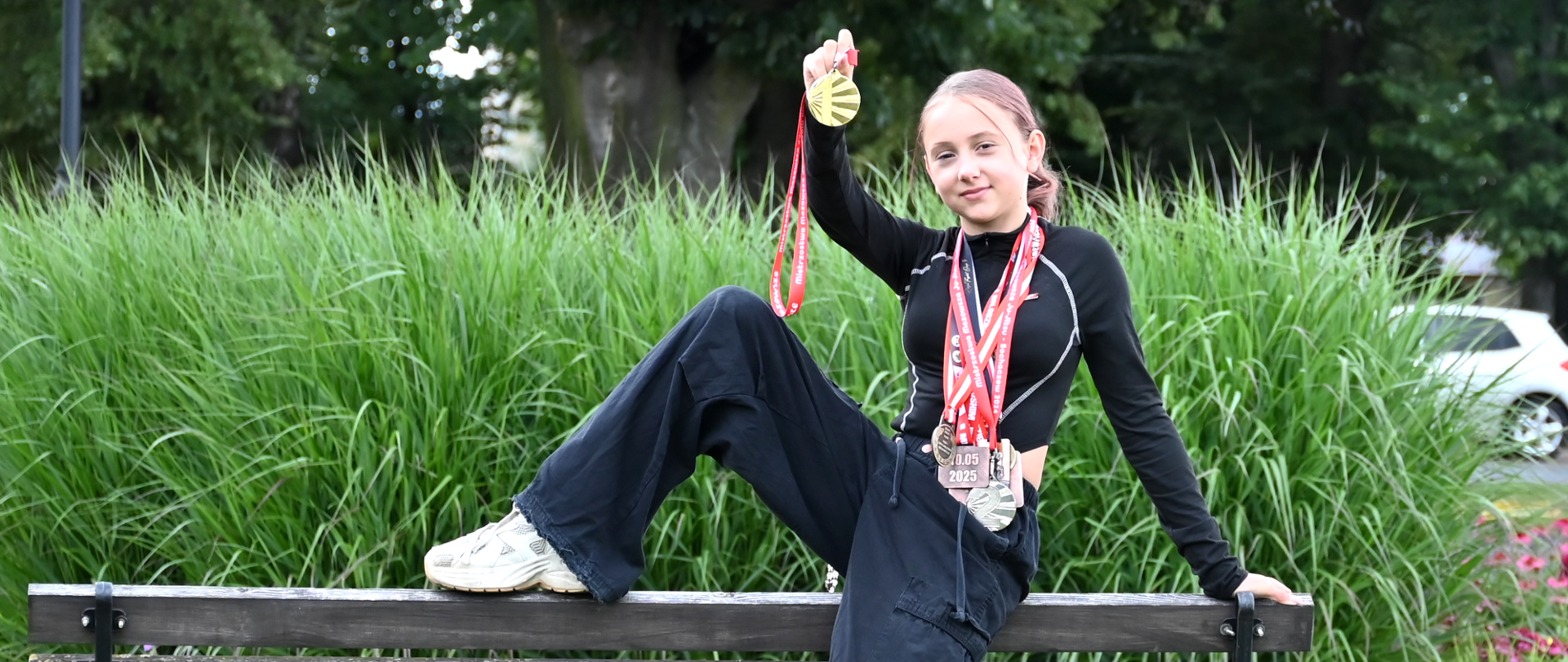 Dziewczynka pozuje na ławce w parku z medalami. Trzyma jedną nogę na oparciu. W tle zieleń i kwiaty.