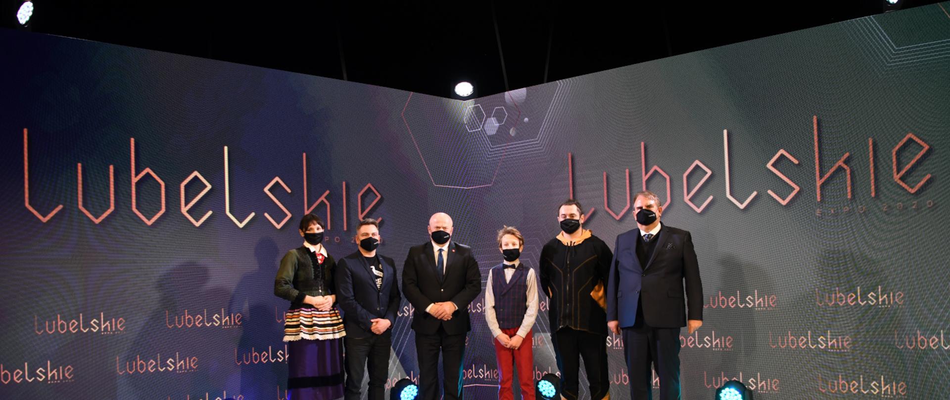 W sali konferencyjnej stoją osoby uczestniczące w konferencji prasowej. W tle widać napis "Lubelskie", który symbolizuje znak województwa lubelskiego, podświetlony przez lampy halogenowe. Wśród uczestników konferencji stoi w miejscu centralnym Marszałek Województwa Lubelskiego Jarosław Stawiarski. 