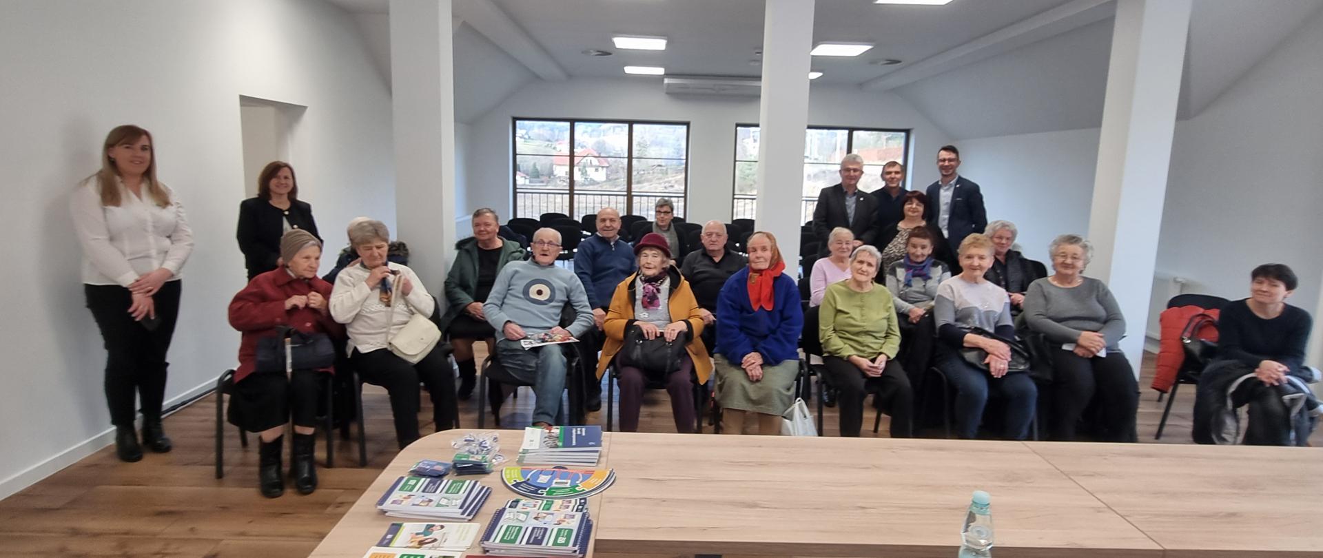 Grupa Seniorów siedzą na krzesłach w sali gdzie odbywa się prezentacja