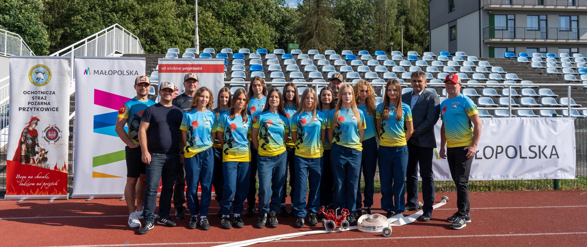 Grupowe zdjęcie Młodzieżowej Drużyny Pożarniczej Dziewcząt OSP Przytkowice wraz z opiekunami i przedstawicielem samorządu. Zawodniczki stoją na bieżni stadionu, ubrane w jednakowe niebiesko-żółte koszulki oraz granatowe spodnie. Przed drużyną leży sprzęt pożarniczy, w tym zwinięte węże i prądownice. W tle widoczne są trybuny, banery OSP Przytkowice i Województwa Małopolskiego oraz zielone drzewa.