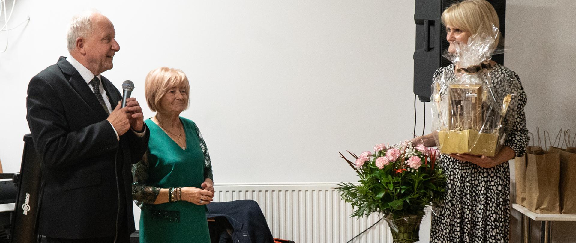 Z lewej strony stoi mężczyzna w czarnym garniturze trzymający mikrofon, obok niego kobieta w zielonej sukience, z prawej strony kobieta stoi trzymając w rękach kosz kwiatów oraz podarunek