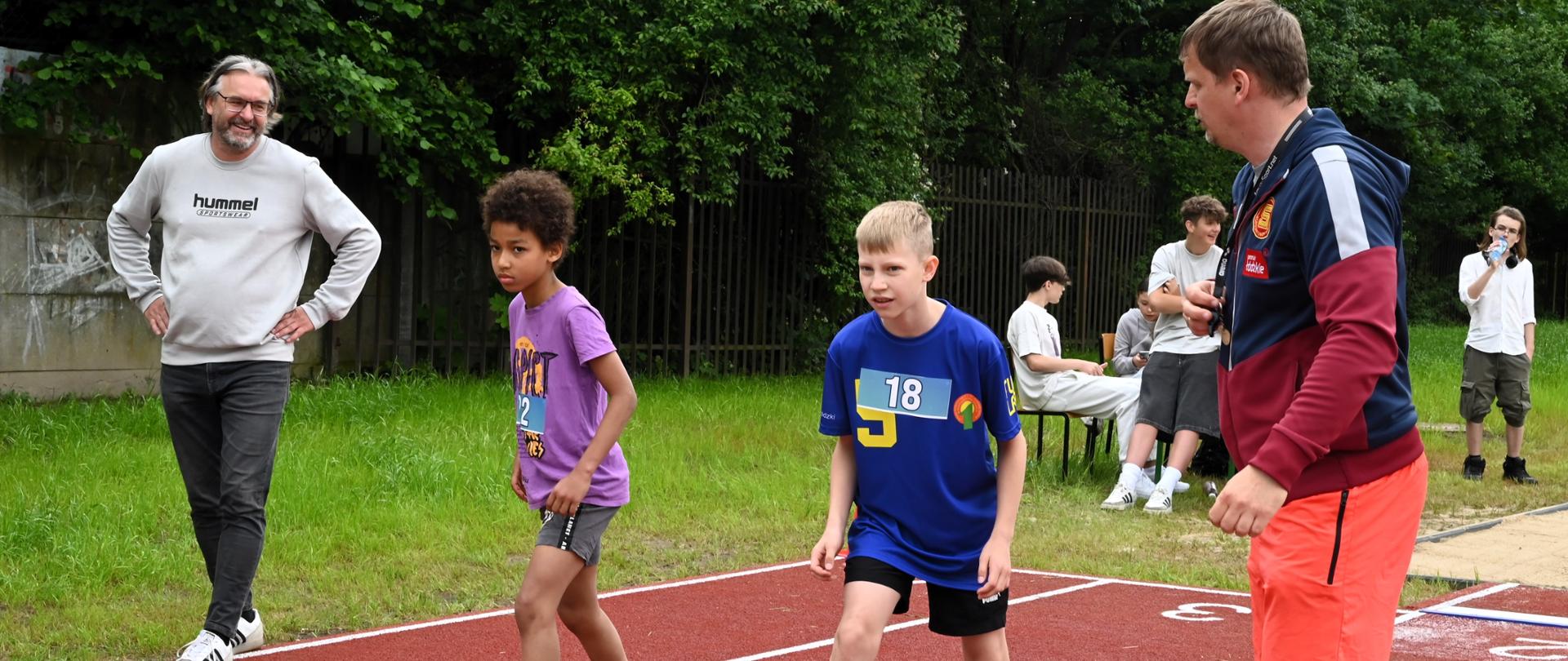 dzieci przygotowujące się do sygnału startowego podczas zawodów w bieganiu. Dwójka chłopców na linii startowej na bieżni. Przy nich stoi sędzia i patrzy czy zgodnie z zasadami wszystko się odbywa. Na drugim planie Stoi Pan Michał Szymczak, czyli współorganizator z ramienia Centrum Sportu i Rekreacji w Konstantynowie Łódzkim