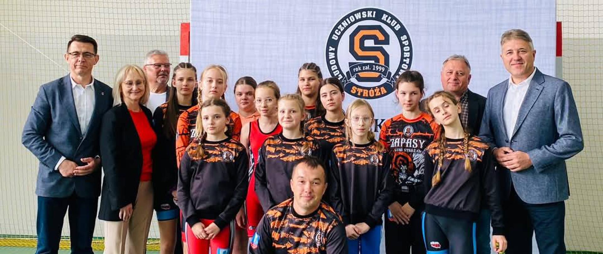 Zawodniczki Ludowego Uczniowskiego Klubu Sportowego Stróża w strojach klubowych na tle białego banera z logiem klubu. Pierwszy od lewej stoi Przewodniczący Rady Powiatu w Kraśniku Jacek Dubiel, zaś pierwszy z prawej Wójt Gminy Kraśnik Artur Domański. Pośrodku przed dziewczynkami klęczy na jedno kolano ich trener klubowy Łukasz Kiełb.