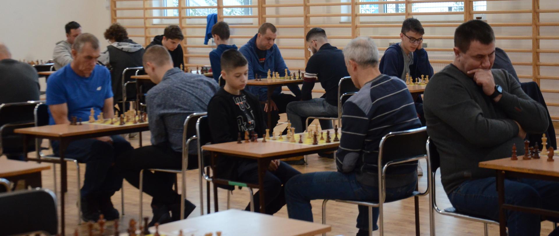 Zawodnicy siedzący na krzesłach podczas pojedynków szachowych przy ustawionych w rzędach stołach na hali sportowej podczas XXIII Otwartego Turnieju Szachowego o Mistrzostwo Powiatu Kraśnickiego w 2023 roku.