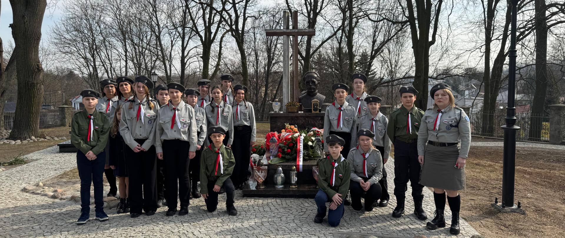 Grupa harcerzy i harcerek w mundurach stoi przed pomnikiem ks. Stanisława Zielińskiego, otoczonym kwiatami i zniczami. W tle widoczny jest park z drzewami, brukowana ścieżka oraz latarnia. Harcerze mają poważne i skupione wyrazy twarzy, oddając hołd pamięci kapłana. 