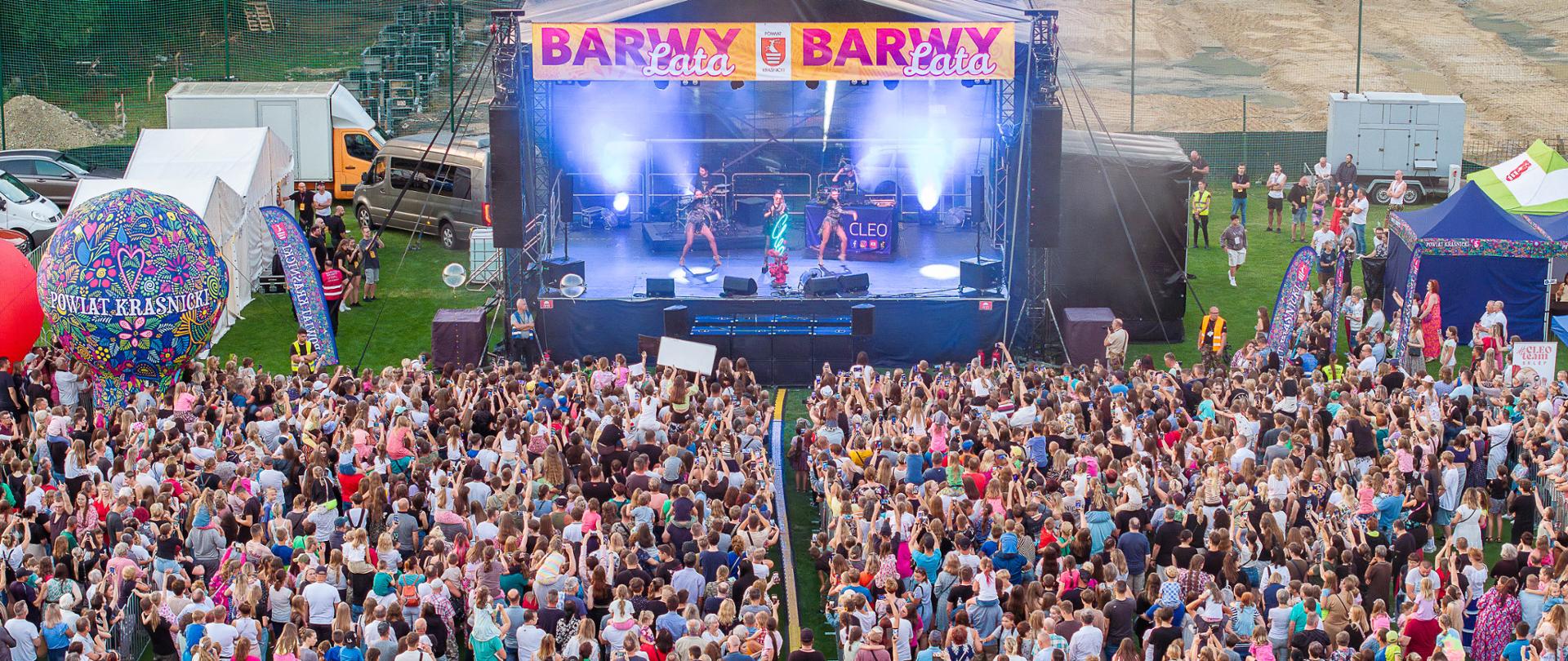 Zdjęcie przedstawia scenę z napisem BARWY LATA, na scenie znajduje się wokalistka i dwie tancerki, przed sceną znajduje się duża grupa ludzi skierowana twarzą w stronę sceny.