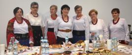 Panie z Koła Gospodyń Wiejskich stoją za stołem z góralskimi tradycyjnymi przysmakami które przygotowały dla przybyłych gości. Na stole widać moskole, pieczywo, sery, ciasto. Panie ubrane w góralskie stroje. 