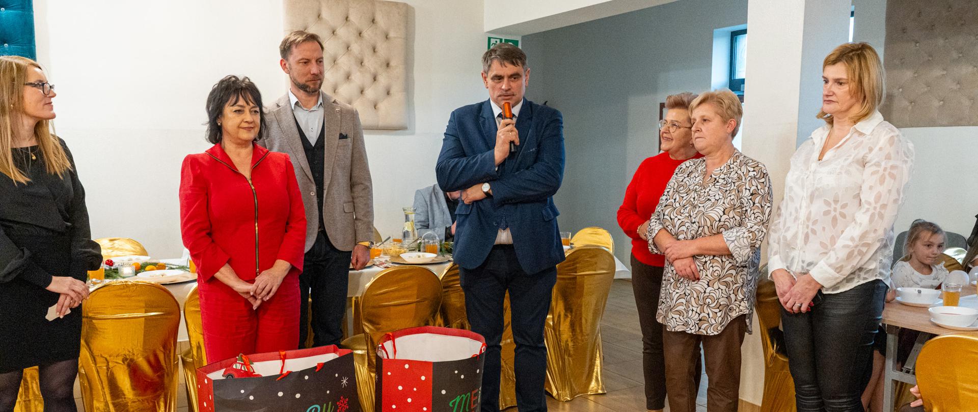 Grupa dorosłych osób stoi w jasnej sali podczas spotkania wigilijnego. W centrum mężczyzna w garniturze przemawia do zebranych, trzymając mikrofon. Obok stoją kobiety i mężczyźni słuchający wystąpienia. Na pierwszym planie widoczne są dwie duże torby z motywem świątecznym, a w tle stoły nakryte do wspólnego posiłku oraz krzesła z złotymi pokrowcami. Wydarzenie ma uroczysty, świąteczny charakter.