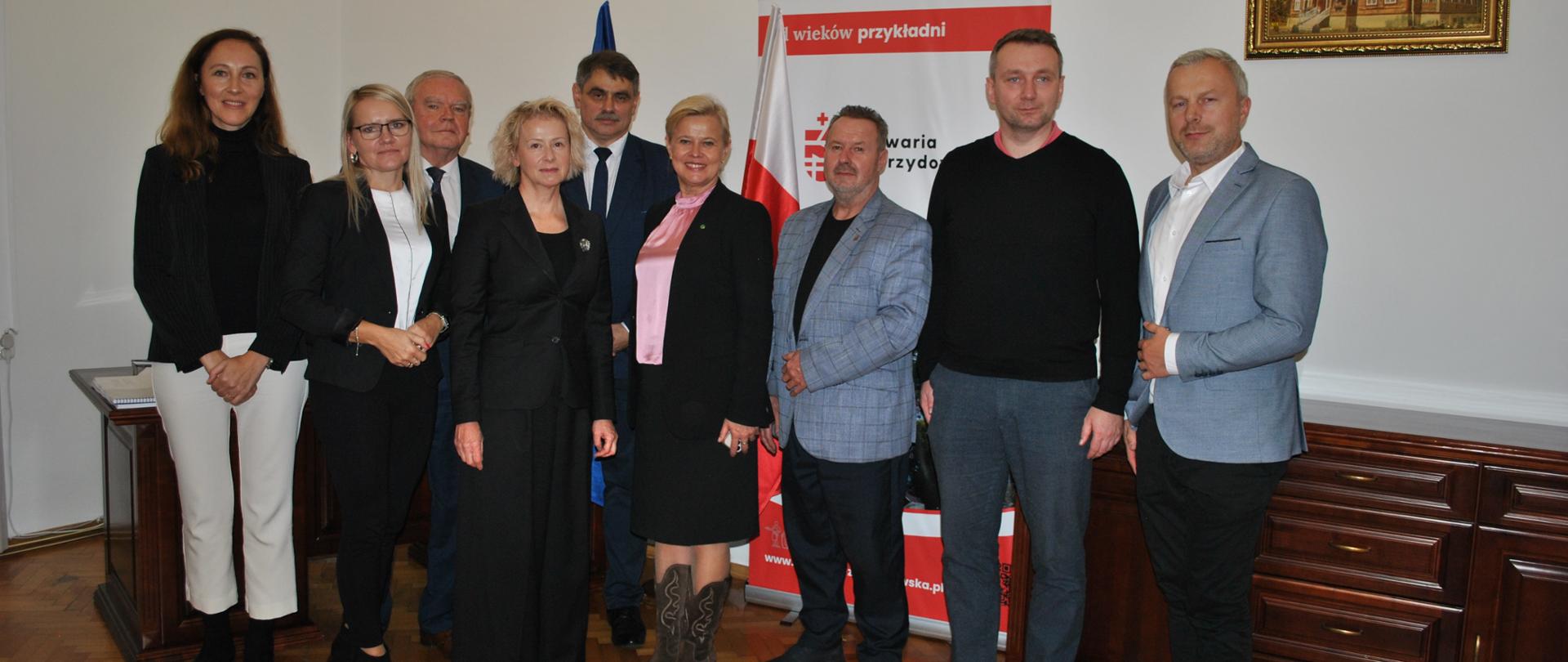 Grupa dziewięciu osób – kobiet i mężczyzn – pozuje wspólnie do zdjęcia w sali urzędu. W tle widać flagę Polski, godło państwowe oraz baner z napisem ‘Od wieków przykładni – Kalwaria Zebrzydowska’. Uczestnicy stoją w rzędzie, ubrani elegancko w stroje formalne, na ścianie widoczny jest również obraz przedstawiający zabytkowy budynek.