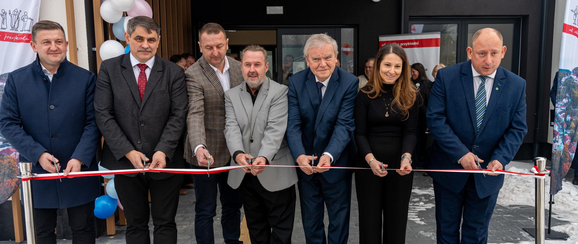 Grupa ośmiu osób stojących przed wejściem do nowoczesnego budynku uczestniczy w uroczystym przecięciu czerwono-białej wstęgi. Wszyscy trzymają nożyczki, ubrani są w eleganckie stroje, a za nimi widoczne są dekoracje z balonów. Wydarzenie odbywa się na zewnątrz, przed obiektem użyteczności publicznej, w zimowej scenerii.