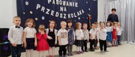Przedszkolaki stoją w sali na tle ścianki "Pasowanie na przedszkolaka"