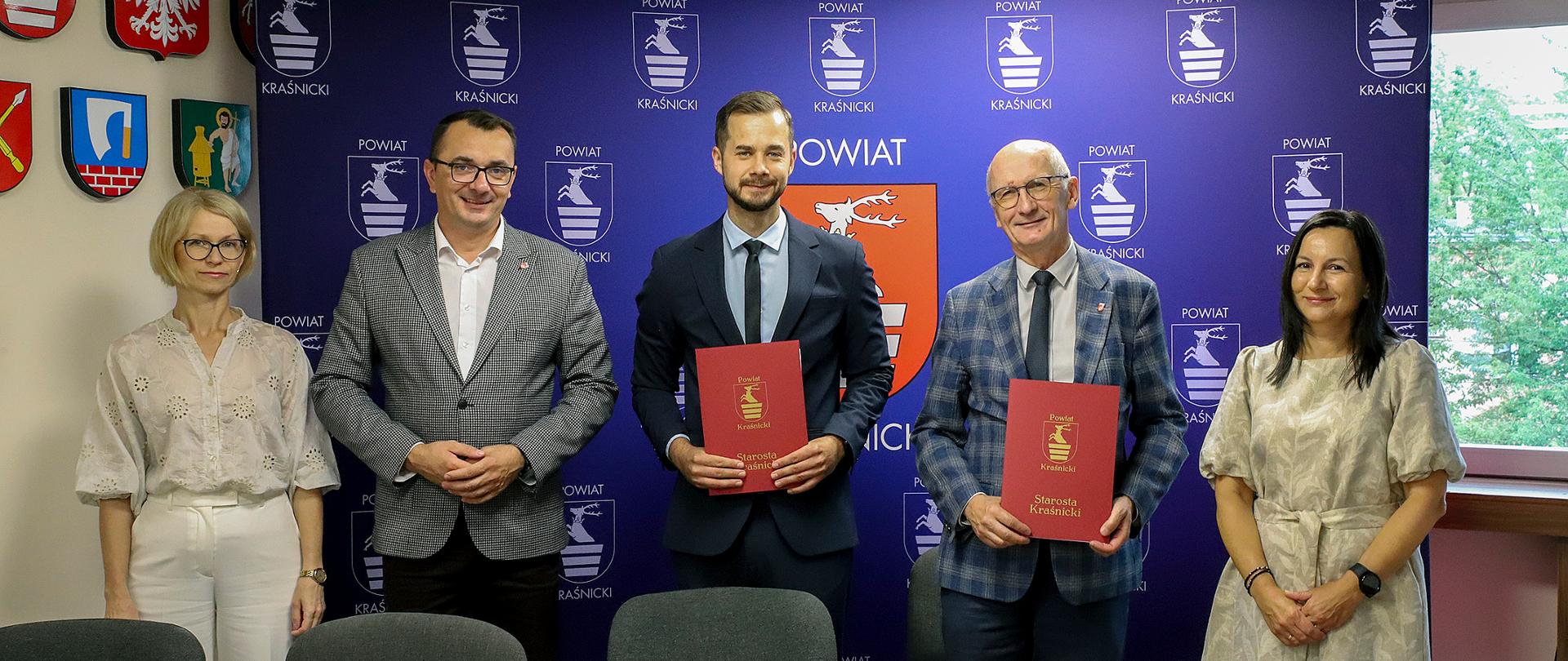 Zdjęcie przedstawia pięć osób (dwie kobiety i trzech mężczyzn) stojących w rzędzie za długim, drewnianym stołem konferencyjnym, na tle niebieskiej ścianki reklamowej Powiatu Kraśnickiego. Na stole leżą dokumenty i długopisy. Dwaj mężczyźni stojący w środku trzymają w dłoniach eleganckie, czerwone teczki z godłem powiatu, co wskazuje na moment po podpisaniu umowy.