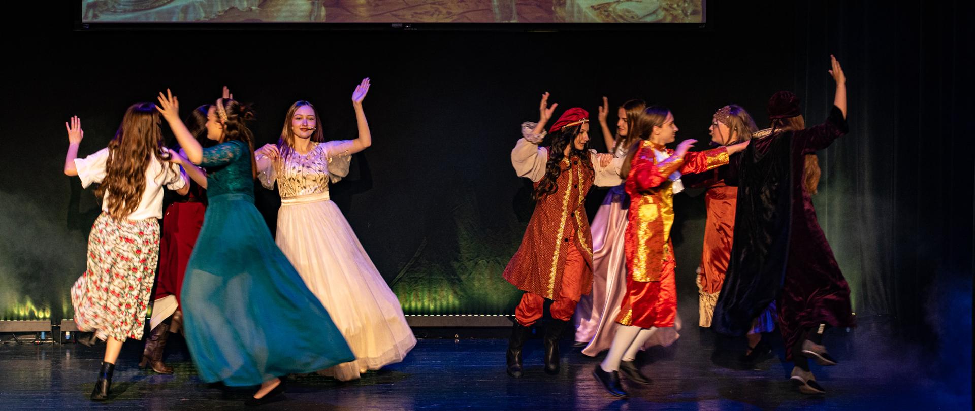 Grupa młodych aktorów w kolorowych kostiumach tanecznych występuje na scenie podczas spektaklu teatralnego. Uczestnicy tworzą krąg, trzymają ręce uniesione do góry i poruszają się w dynamicznym tańcu. W tle widoczna jest projekcja eleganckiej, bogato udekorowanej sali bankietowej z kwiatami i świecami. Scena oświetlona jest ciepłym światłem, a całość tworzy radosną, bajkową atmosferę przedstawienia.