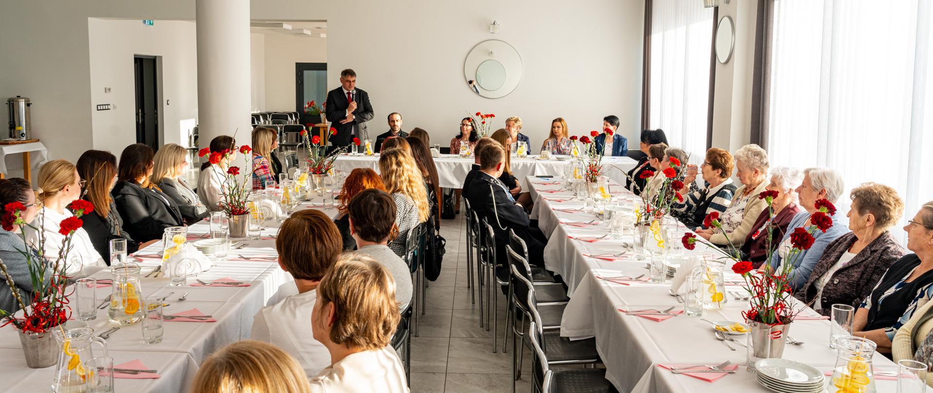 Sala w remizie OSP w Leńczach podczas walnego zebrania Koła Gospodyń Wiejskich – uczestnicy siedzą przy długich stołach udekorowanych czerwonymi goździkami, a mężczyzna przemawia do zgromadzonych.
