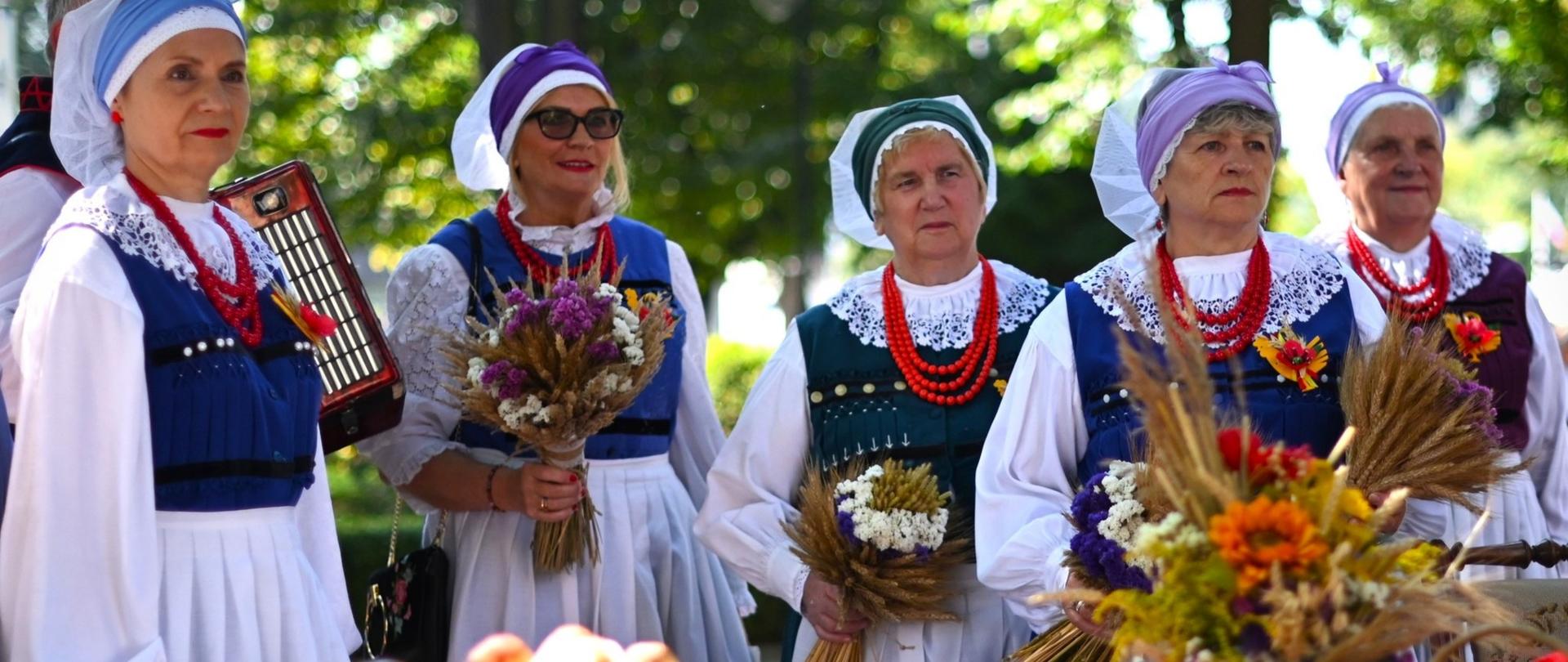 Kobiety w strojach ludowych, w tle zielona drzewa. Z przodu pieczywo, tradycyjna wiązanka ze zbóż i kwiatów.