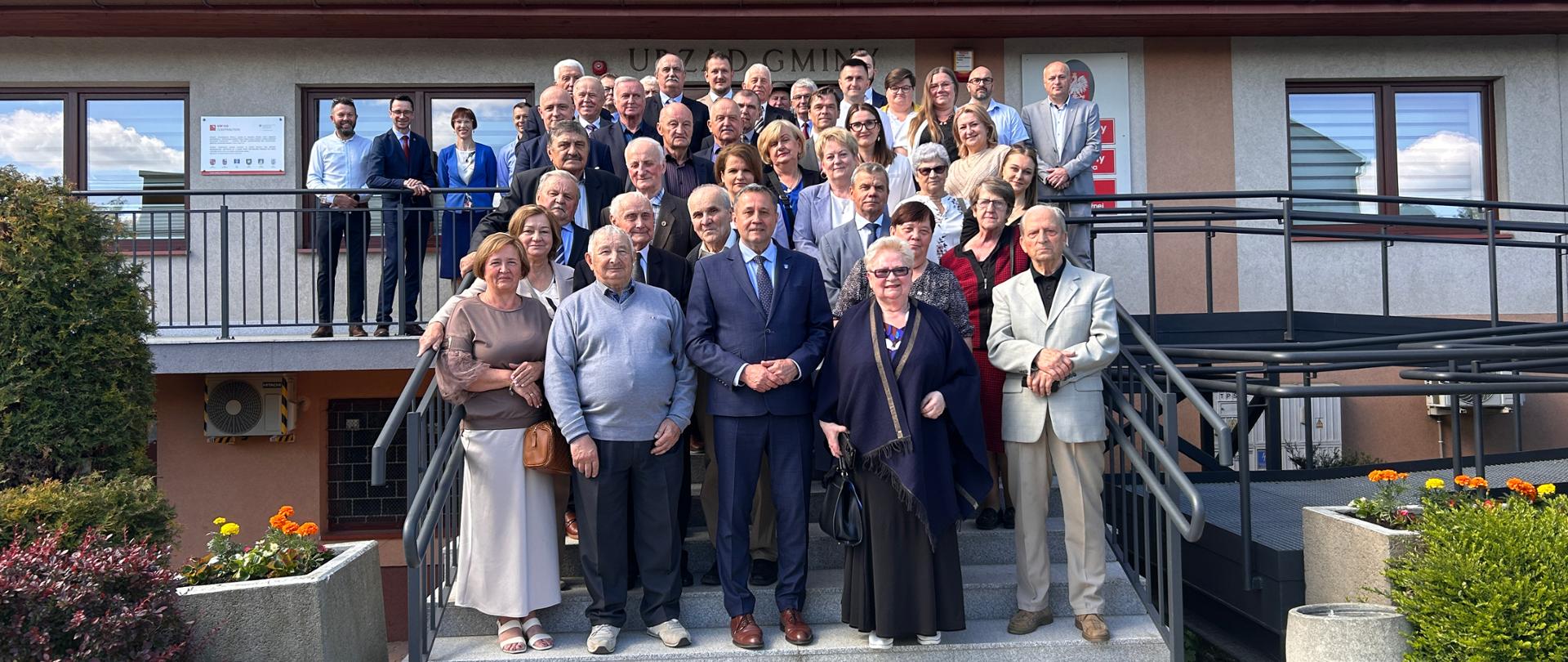 Grupa osób pozuje do wspólnego zdjęcia na schodach przed budynkiem Urzędu Gminy.

