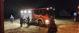 Czerwony samochód straży pożarnej nocą. Odgrodzony taśmą i oświetlony reflektorami z górnej części samochodu. Przy samochodzie stoją strażach i osoba odwrócona plecami.