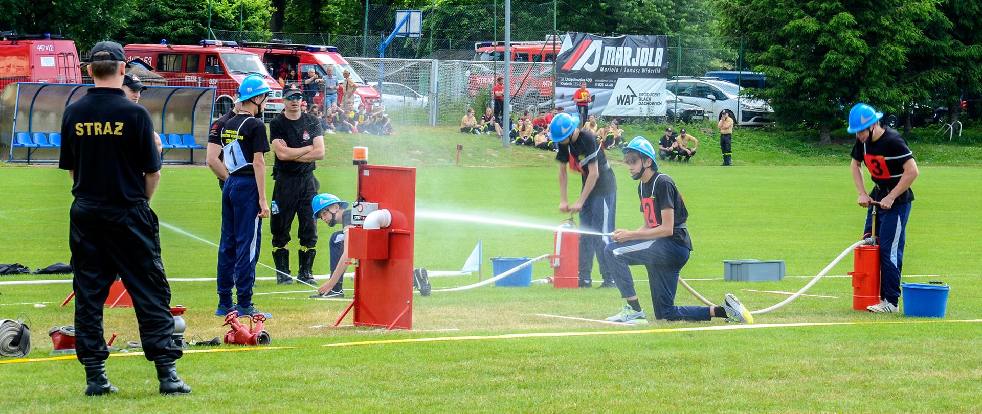 Zdjęcie przedstawia młodych strażaków, którzy na przygotowanym torze biorą udział w zawodach. Dwie pary zawodników kierują strumień wody do celu. Zmagania obserwowane są przez sędziów. Zawody rozgrywają się na murawie stadionu sportowego. W tle znajdują się zaparkowane wozy strażackie.