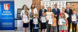 Na zdjęciu znajduje się grupa dzieci prezentująca dyplomy. Z tyłu za nimi na tle ścianki Gminnego Ośrodka Kultury stoją członkowie jury i organizatorzy. Po lewej stronie stoi rollup Powiatu Kraśnickiego