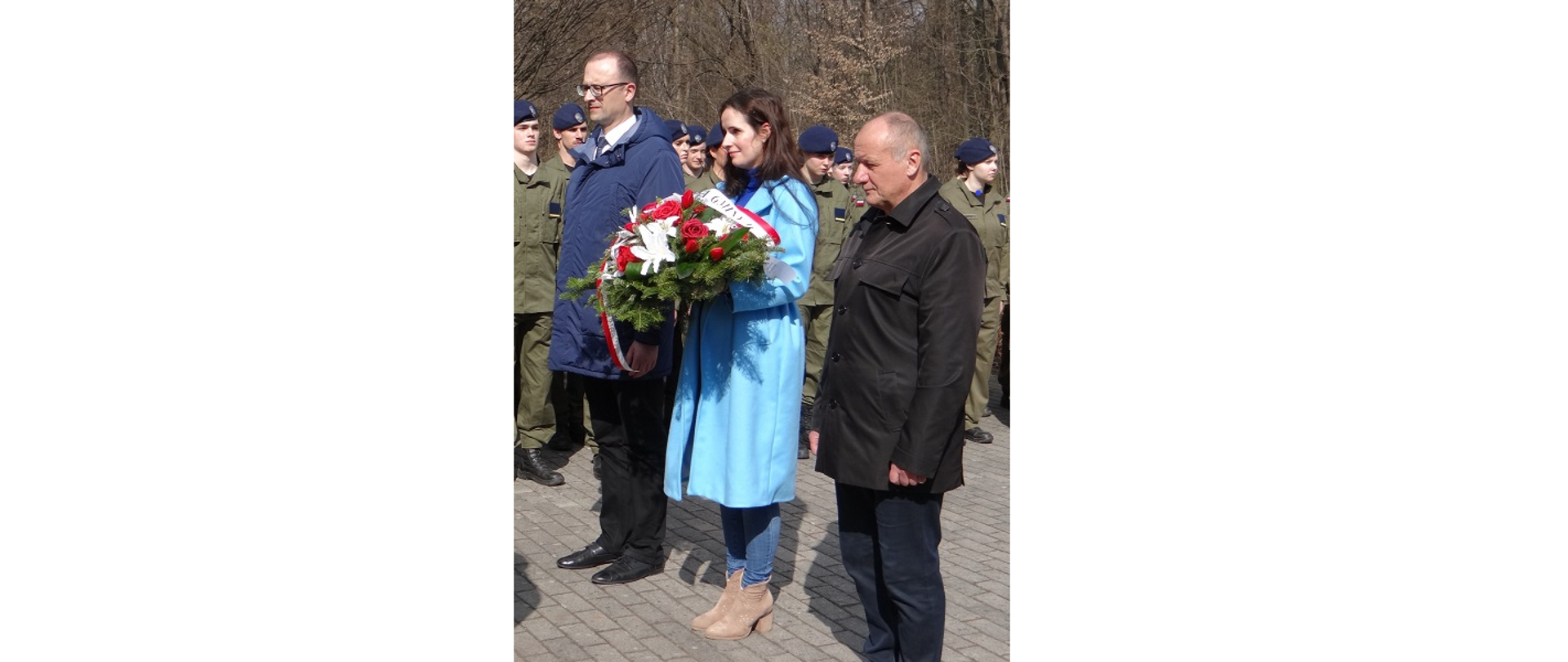 Delegacja z Gminy Hażlach składa pod pomnikiem wiązankę kwiatów. Na pierwszym planie trzy osoby z delegacji. Za nimi młodzi ludzie w mundurach