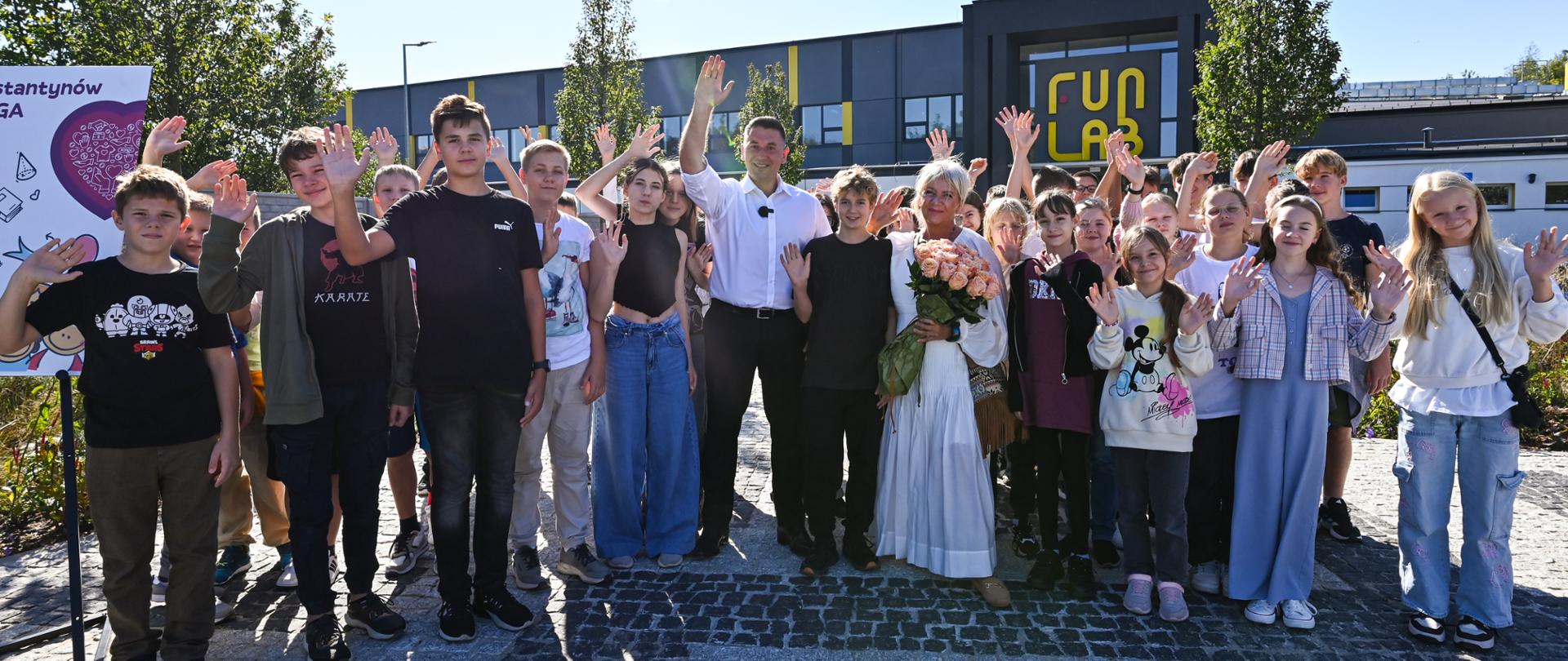 Zdjęcie grupowe uczniów przed szkołą, słoneczna pogoda, machają do widzów.