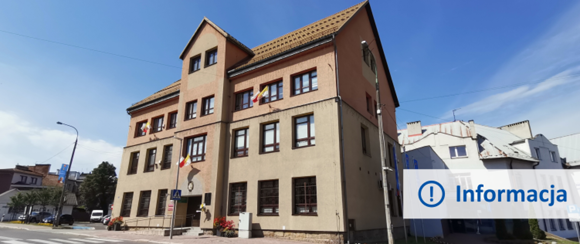 Budynek Urzędu Miasta Kalwarii Zebrzydowskiej w słoneczny dzień. Po lewej parking z widoczną latarnią. Po prawej fragment budynku Policji.