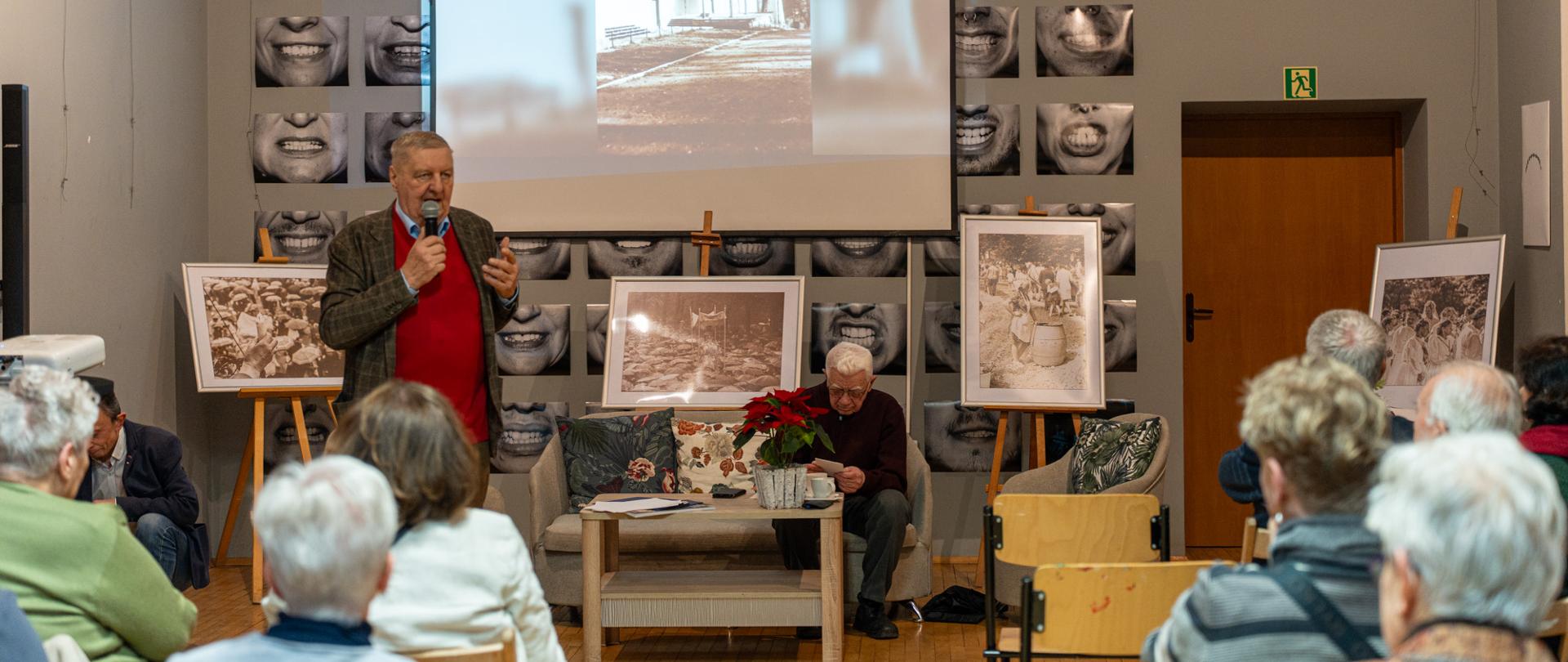 Spotkanie autorskie w sali z publicznością siedzącą na krzesłach. Na pierwszym planie mężczyzna stoi i przemawia do zgromadzonych, trzymając mikrofon. Obok, przy stoliku, siedzi drugi mężczyzna przeglądający dokumenty. Za nimi widoczny jest ekran z wyświetloną archiwalną fotografią zabudowań oraz ekspozycja czarno-białych zdjęć na sztalugach. Wydarzenie ma charakter kulturalny i historyczny, a uczestnikami są głównie osoby starsze.