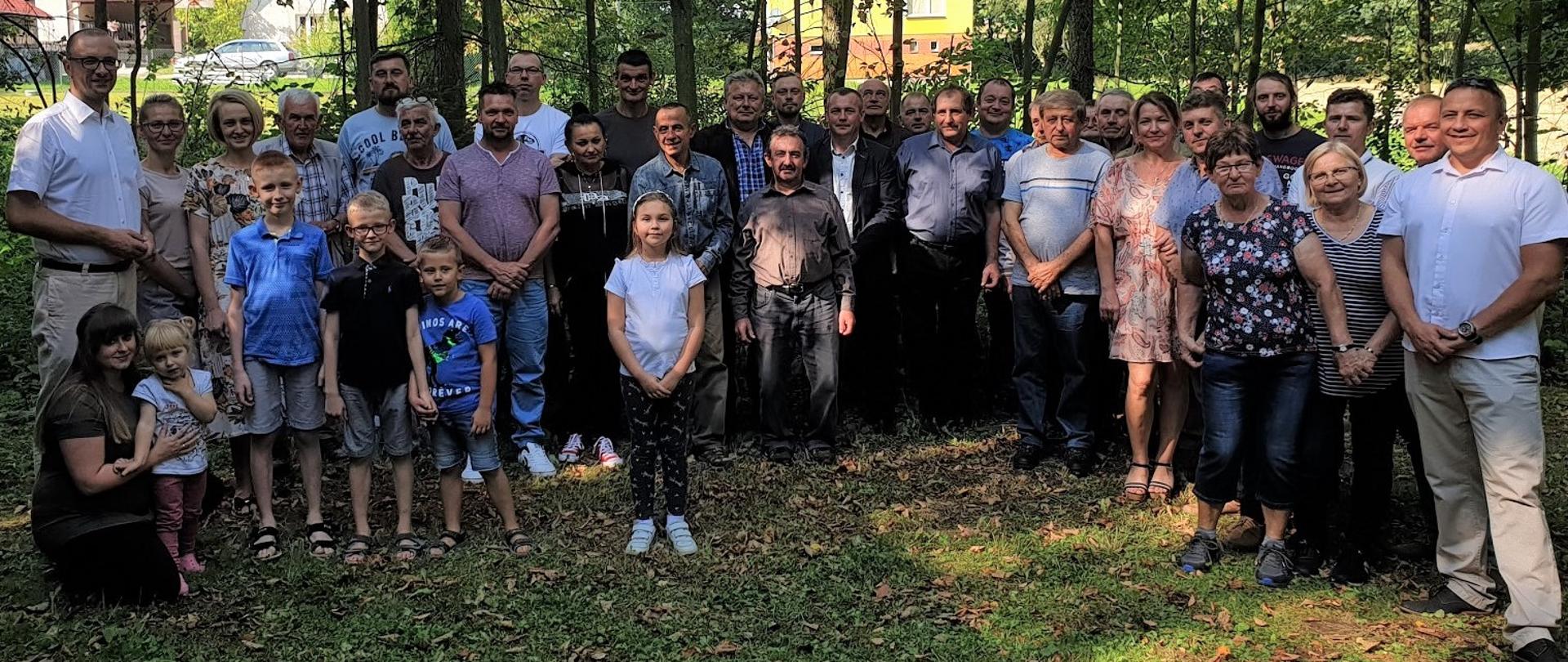 Pamiątkowa fotografia uczestników spotkania. Zebrane osoby, w tym kilkoro dzieci, pozują ustawieni w podkowę na polanie. Wokół zadrzewiony teren pasieki. 