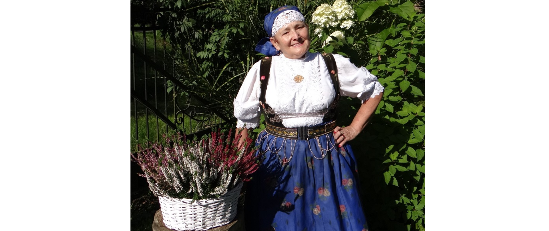 Maria Żyła, przewodnicząca Koła Gospodyń Wiejskich w Kończycach Wielkich. Kobieta w stroju cieszyńskim pozuje do fotografii w ogrodzie na tle roślinności 