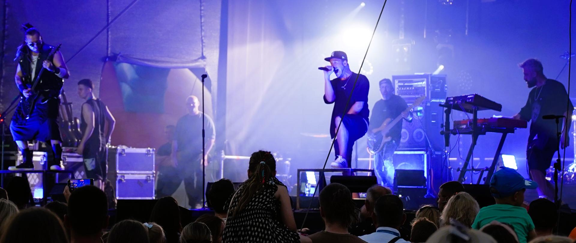 Na zdjęciu widać 6 mężczyzn znajdujących się na scenie, którzy gra koncert. Przed sceną znajduje się liczna publiczność.
