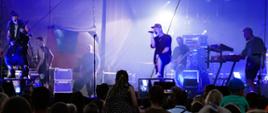 Na zdjęciu widać 6 mężczyzn znajdujących się na scenie, którzy gra koncert. Przed sceną znajduje się liczna publiczność.