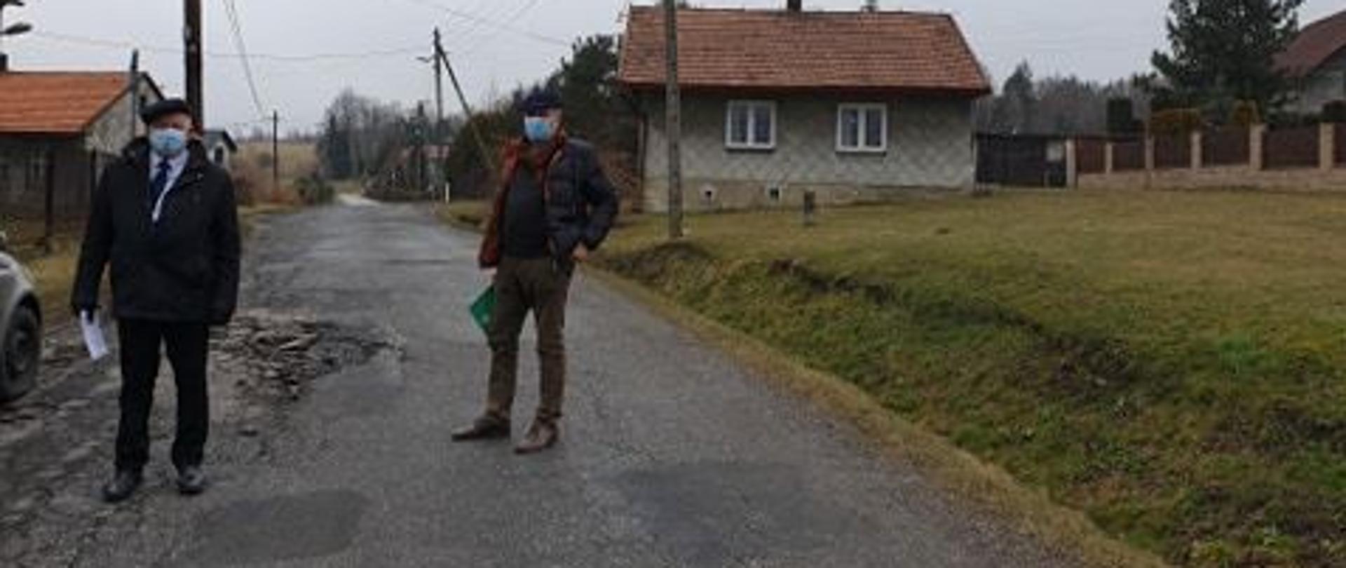 Zdjęcie uszkodzonej drogi we wsi Podolany. Na pierwszym planie wizytujący wraz z pracownikiem urzędu Burmistrz miasta Kalwaria Zebrzydowska. W oddali po obu stronie drogi budynki.