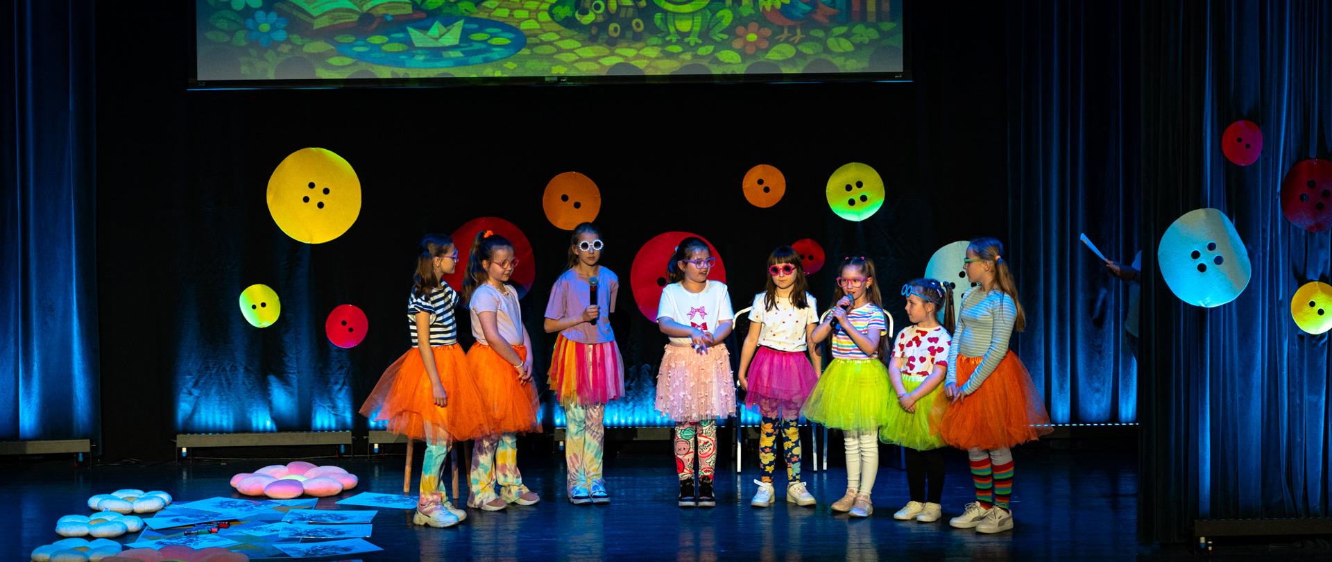 Na scenie teatralnej stoi grupa dzieci w kolorowych, bajkowych strojach – spódniczkach tutu i wzorzystych ubraniach. Kilkoro z nich trzyma mikrofony, wygląda jakby śpiewały lub mówiły podczas występu. W tle widoczna jest duża, barwna ilustracja przedstawiająca fantastyczny świat z zamkiem, tęczą, roślinami i zwierzętami, nawiązująca do bajkowej scenografii. Dekorację uzupełniają kolorowe, okrągłe elementy przypominające guziki rozmieszczone na kurtynie. Atmosfera jest radosna i teatralna, sugerująca występ dziecięcy lub przedstawienie.