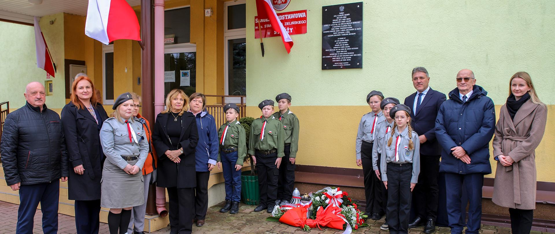 Pamiątkowe zdjęcie przy tablicy znajdującej się na budynku szkoły. Zdjęcie przedstawia grupę ludzi zgromadzonych wokół tablicy, przy której złożono biało-czerowne kwiaty i znicze. Przy tablic wartę pełnią młodzi harcerze. W tle na budynku szkoły znajdują się biało-czerwone flagi.