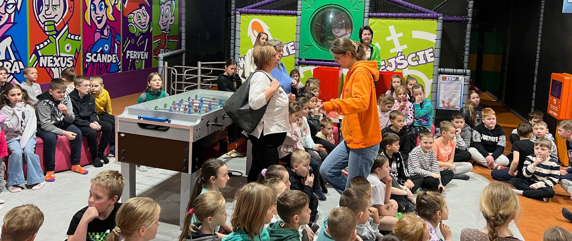 Grupa dzieci siedzących na podłodze w kolorowej sali zabaw podczas ferii zimowych. W tle widoczne są konstrukcje do zabawy, grafiki na ścianach oraz piłkarzyki. Dzieci uważnie słuchają opiekunów stojących pośrodku sali, a atmosfera spotkania ma charakter integracyjny i zorganizowany.